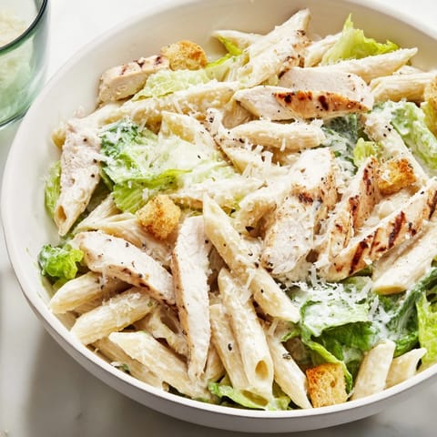 A Caesar Pasta Chicken Bowl garnished with shaved Parmesan and halved cherry tomatoes, served warm on a dinner table.